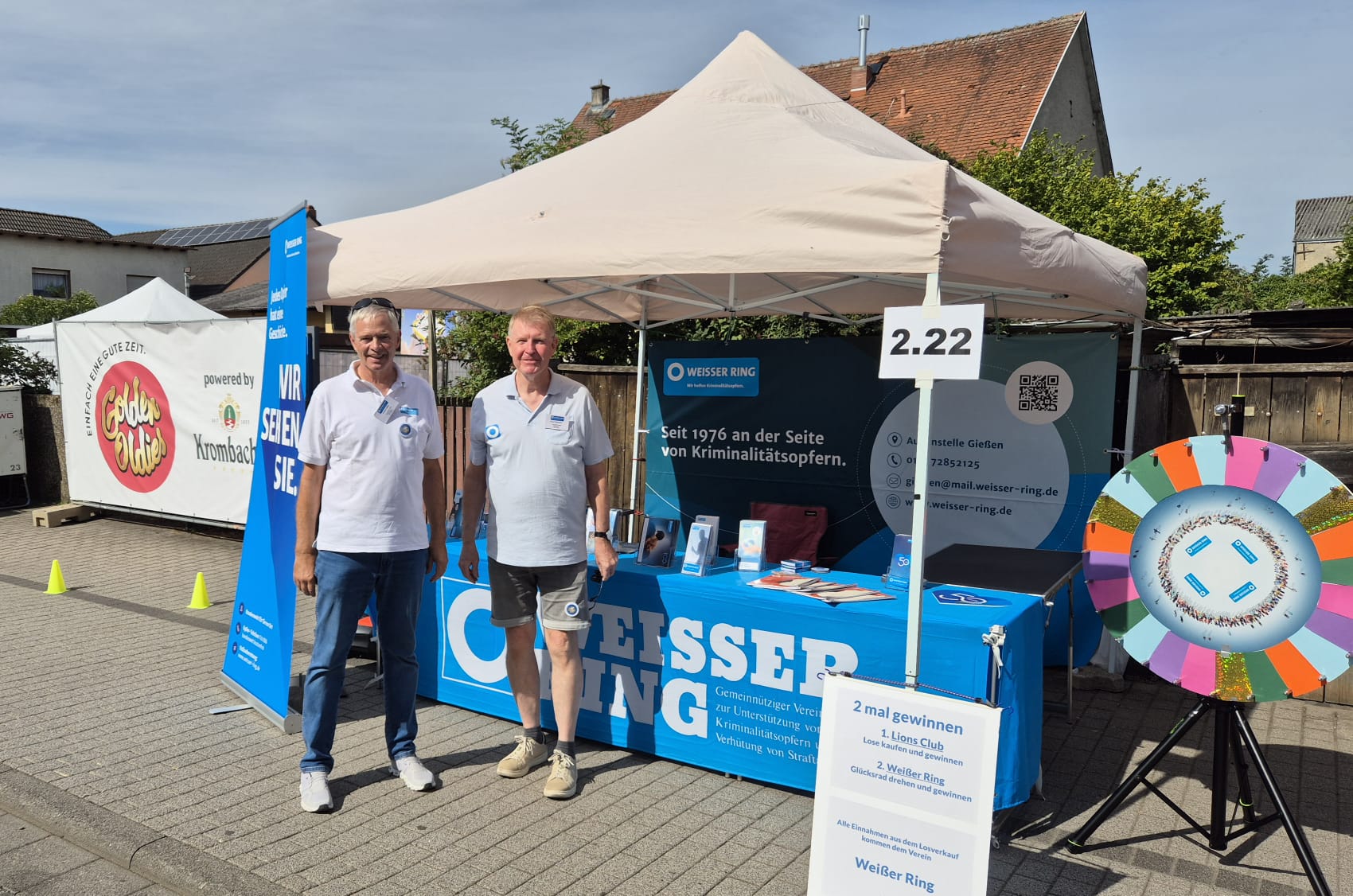 Zwei Mitarbeiter des WEISSEN RINGS vor einem Pavillon, daneben ein Glücksrad