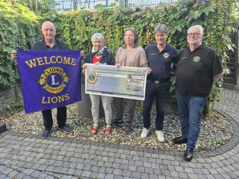 Auf dem Bild sind fünf Menschen zu sehen. Zwei Frauen und drei Männer. Ein Mann hält eine Flagge des Lions Clubs, die beiden Frauen halten gemeinsam ein Spendenscheck.