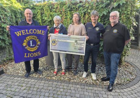 Auf dem Bild sind fünf Menschen zu sehen. Zwei Frauen und drei Männer. Ein Mann hält eine Flagge des Lions Clubs, die beiden Frauen halten gemeinsam ein Spendenscheck.