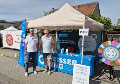 Zwei Mitarbeiter des WEISSEN RINGS vor einem Pavillon, daneben ein Glücksrad
